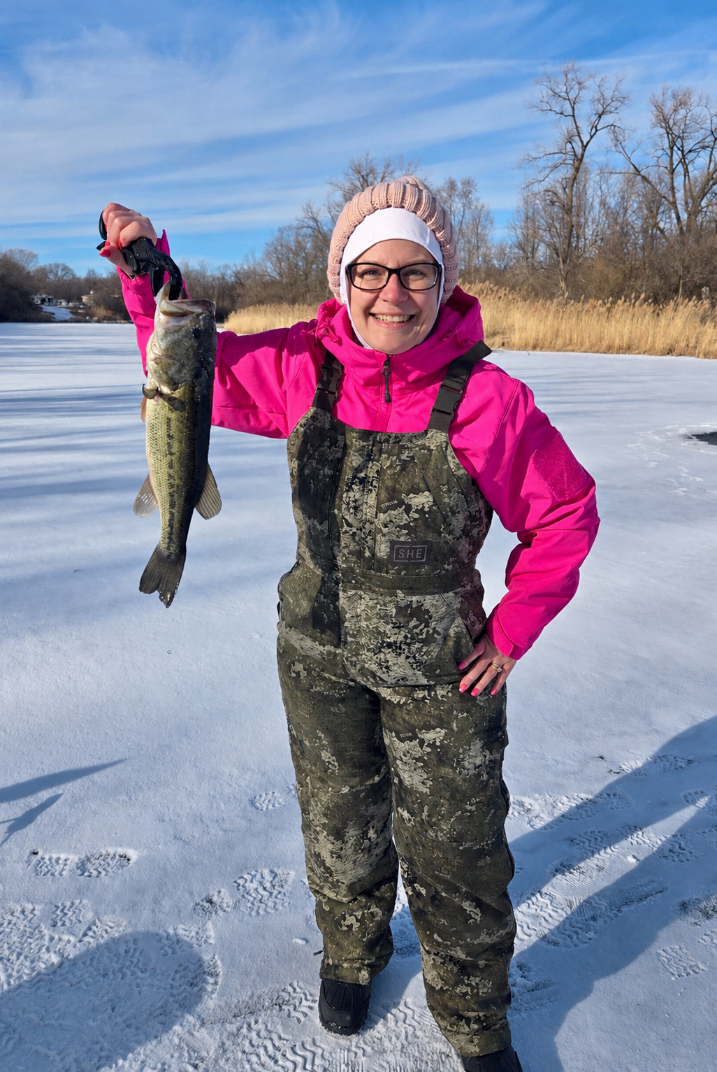 Female Deer Creek member in camo bibs holding a bass caught ice fishing on a frozen lake in Illinois